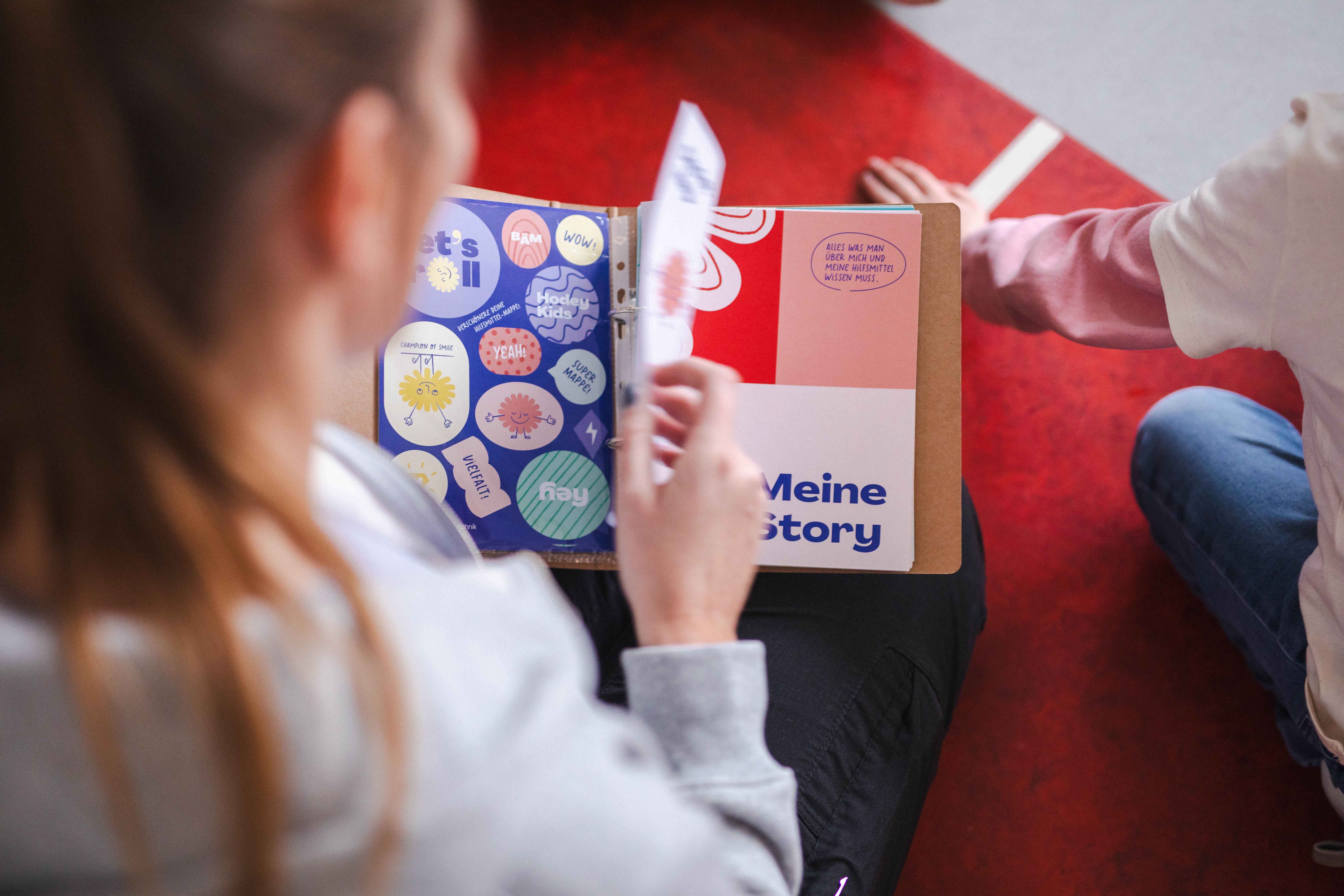 Blick über die Schulter einer Hodey Kids Mitarbeiterin. Sie blättert durch den Hilfsmittelpass. Neben ihr sitzt ein Kind.
